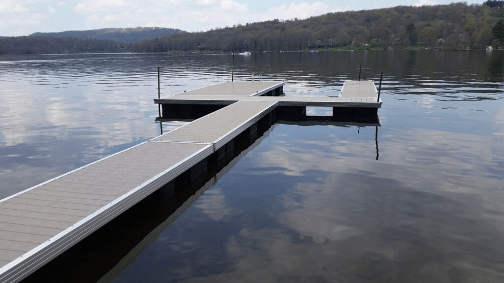 A wooden dock with multiple walkways extends over a calm lake, surrounded by distant trees and hills under a cloudy sky.