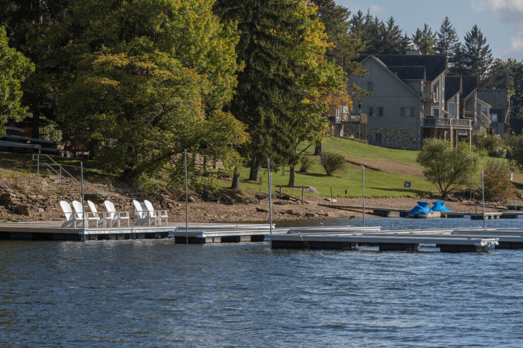A lakeside dock with white chairs and nearby houses, surrounded by trees and grassy areas under a partly cloudy sky.