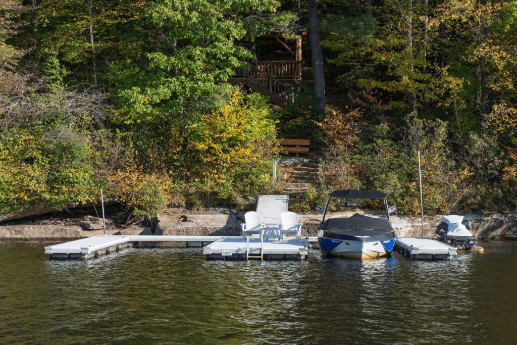 A lakeside dock with two lounge chairs, a covered boat, and a jet ski, backed by trees and a wooden staircase.