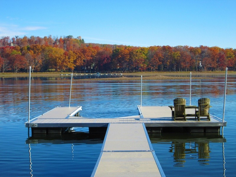 A dock with two chairs overlooks a calm lake surrounded by trees with autumn foliage under a clear sky.