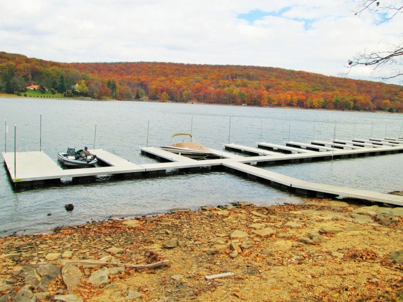 A dock with boats sits on a lake, surrounded by autumn trees and a rocky shoreline under a partly cloudy sky.