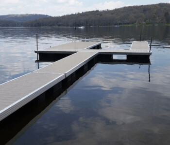 A wooden dock with multiple walkways extends over a calm lake, surrounded by distant trees and hills under a cloudy sky.