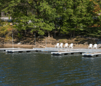 Floating dock with several white chairs, calm water in foreground, and trees lining the shore in the background.