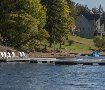 A lakeside dock with white chairs and nearby houses, surrounded by trees and grassy areas under a partly cloudy sky.