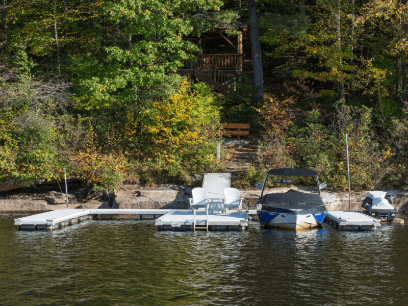 A lakeside dock with two lounge chairs, a covered boat, and a jet ski, backed by trees and a wooden staircase.
