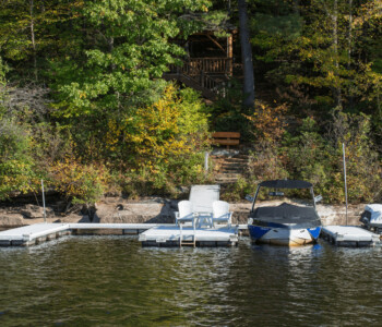 A lakeside dock with two lounge chairs, a covered boat, and a jet ski, backed by trees and a wooden staircase.