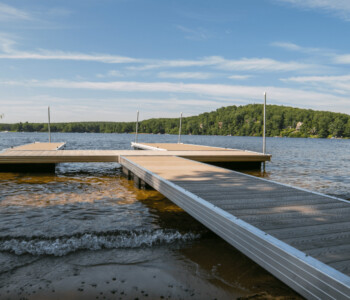 A wooden dock extends into a calm lake bordered by trees under a blue sky with scattered clouds.