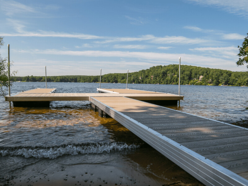 A wooden dock extends into a calm lake bordered by trees under a blue sky with scattered clouds.