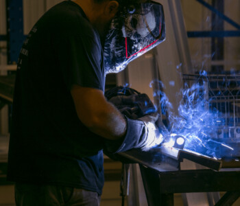 Person wearing protective gear welding metal in a workshop, with sparks and blue light visible.